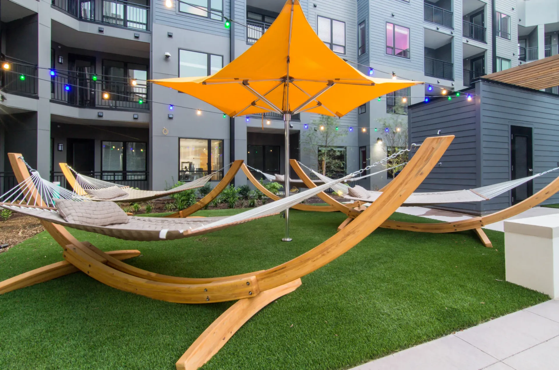 Outdoor community courtyard with curved wooden hammocks and string lights under an orange canopy.