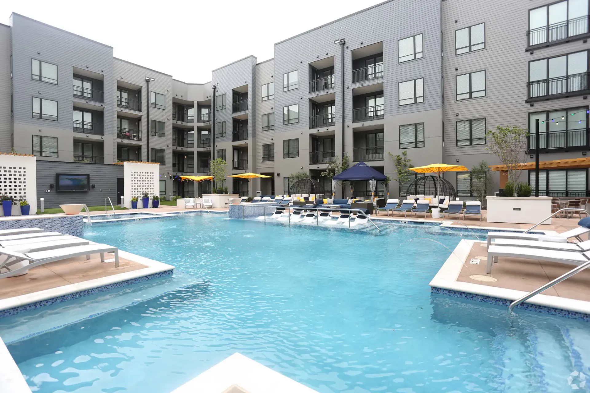 Swimming pool in front of a modern apartment complex with multiple stories, loungers, and blue umbrellas.