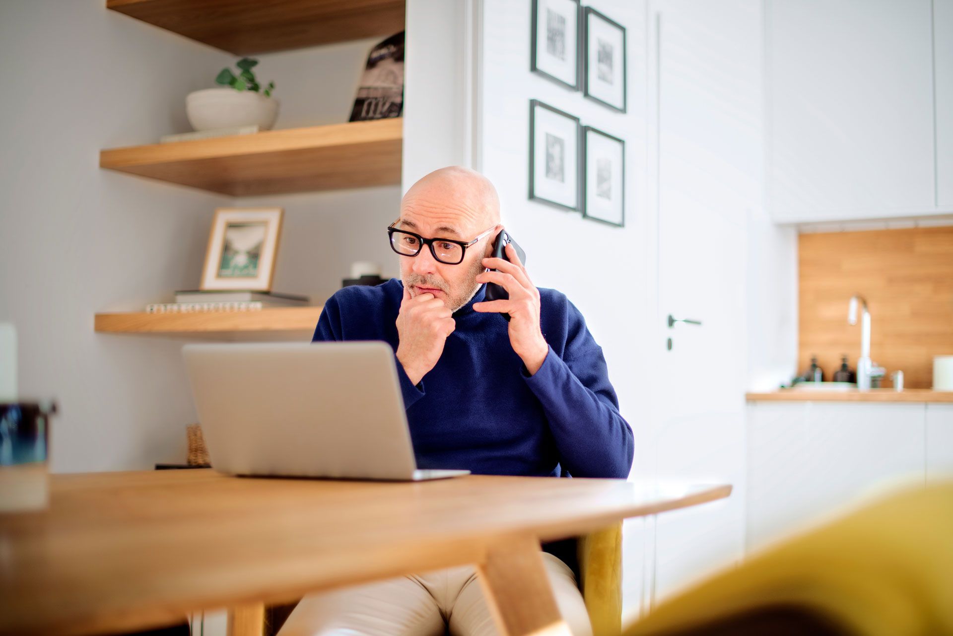 Man on phone looking at laptop in a home office setting.