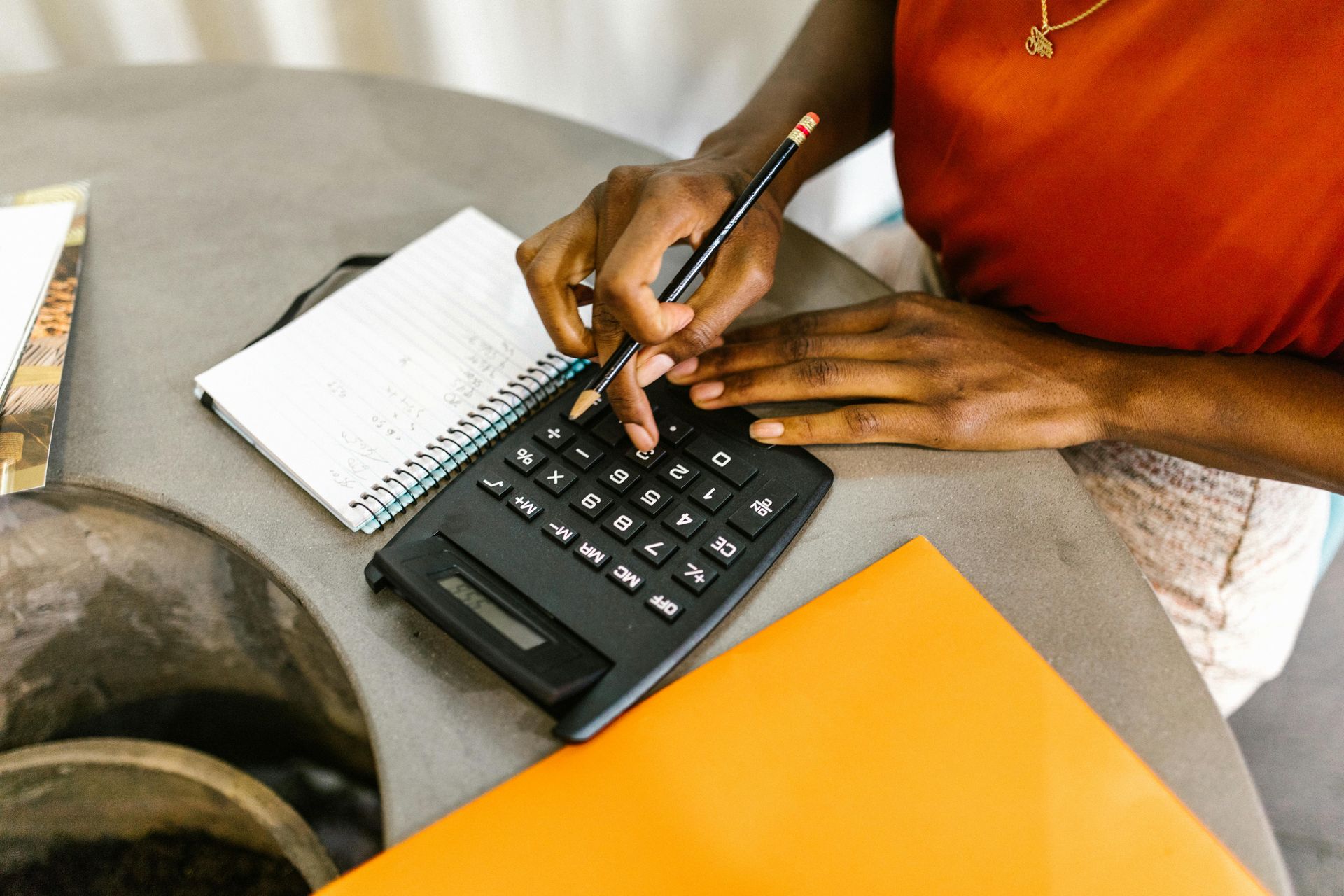 Person using a calculator and notepad on a table, orange top, pencil in hand.