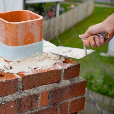A person uses a trowel to apply mortar around a clay chimney flue on top of a brick chimney.