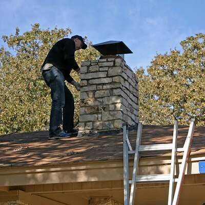 A person wearing dark clothing repairs a stone chimney on a residential roof near a ladder under a clear blue sky.