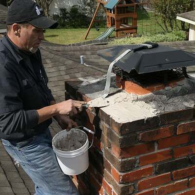 Worker applying mortar to a brick chimney outdoors, with a bucket and trowel.
