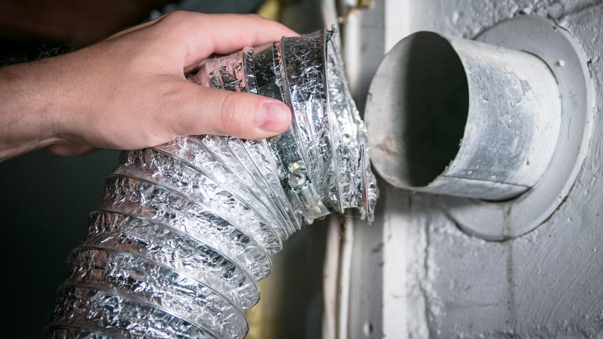 A hand holds a flexible metallic dryer vent hose near a wall exhaust pipe.
