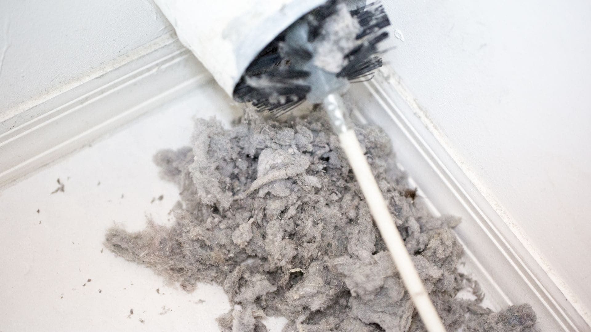 A dryer vent cleaning brush pulls a large pile of grey lint out of a white wall vent.