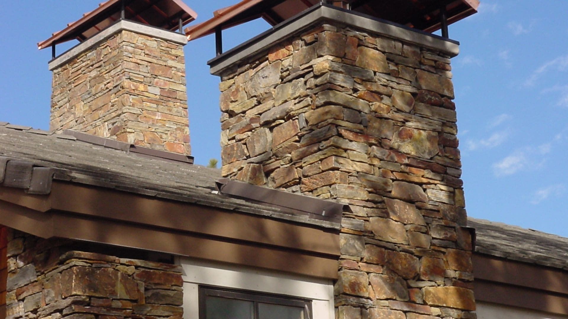 Two stone chimneys with metal caps rise from a house roof against a blue sky.