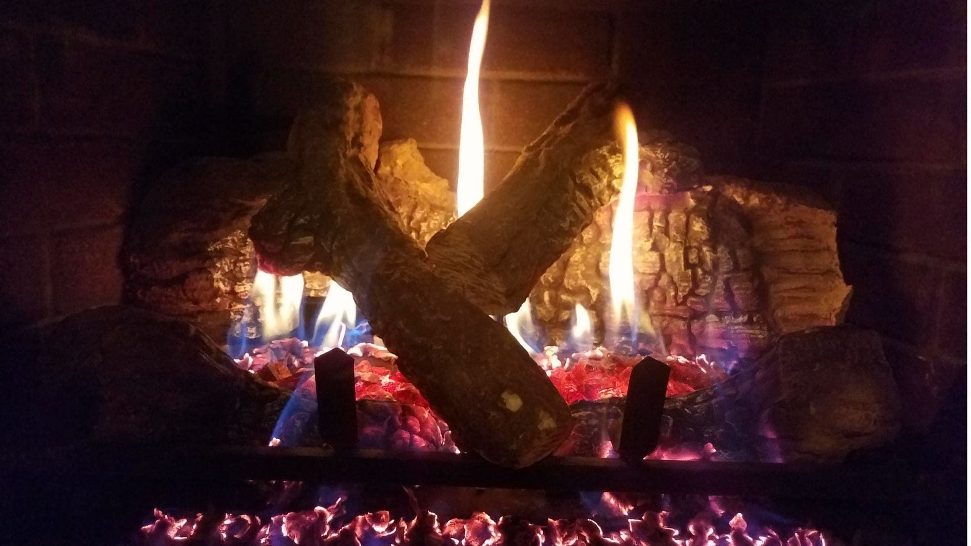 A gas fireplace with glowing, artificial logs and bright orange and blue flames against a brick background.