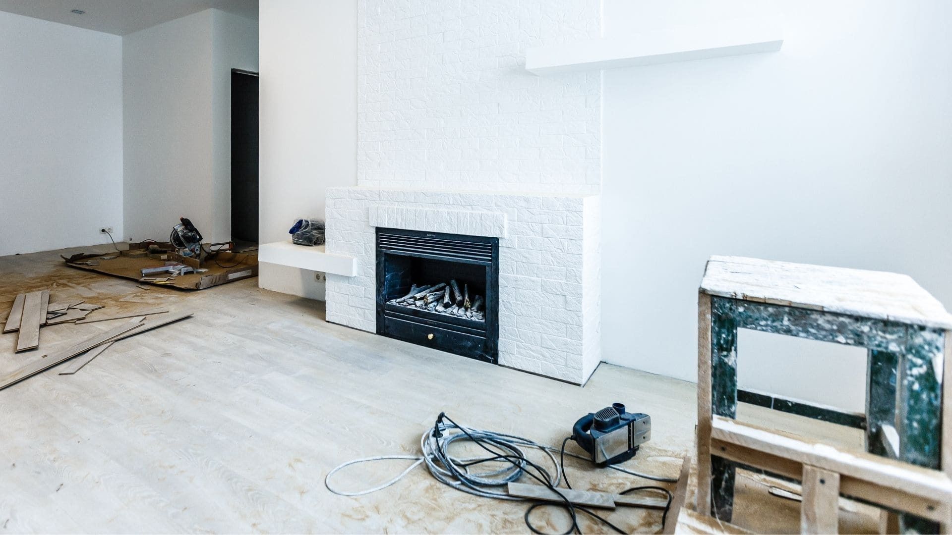 A white living room under renovation with a brick fireplace, a wall-mounted shelf, construction tools, and debris.