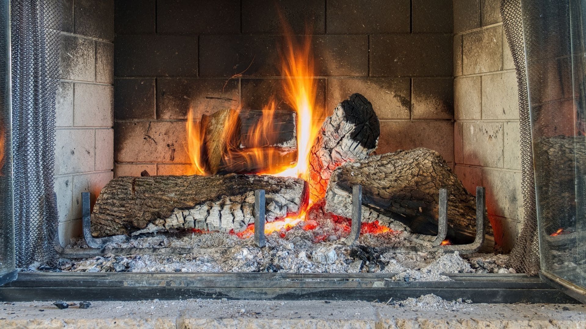 A fire burns with bright orange flames between two logs inside a stone fireplace lined with gray ash.