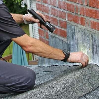 A person wearing gloves inspects roof flashing along a brick wall using a flashlight.