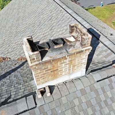 An aerial view of a tan brick chimney on a shingled roof, showing three open, dark flue openings at the top.