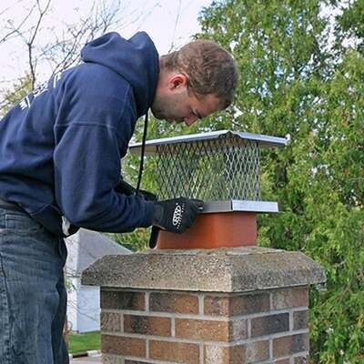 A person wearing a blue hoodie and gloves installs a metal chimney cap onto a brick chimney on a roof.