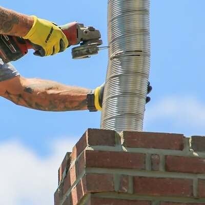 A worker wearing yellow gloves uses a power cutter to trim a metal chimney liner atop a brick chimney against a blue sky.