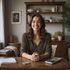Woman seated at a wooden desk smiling. Bookshelf and plants in the background. Documents and a calculator on the desk.