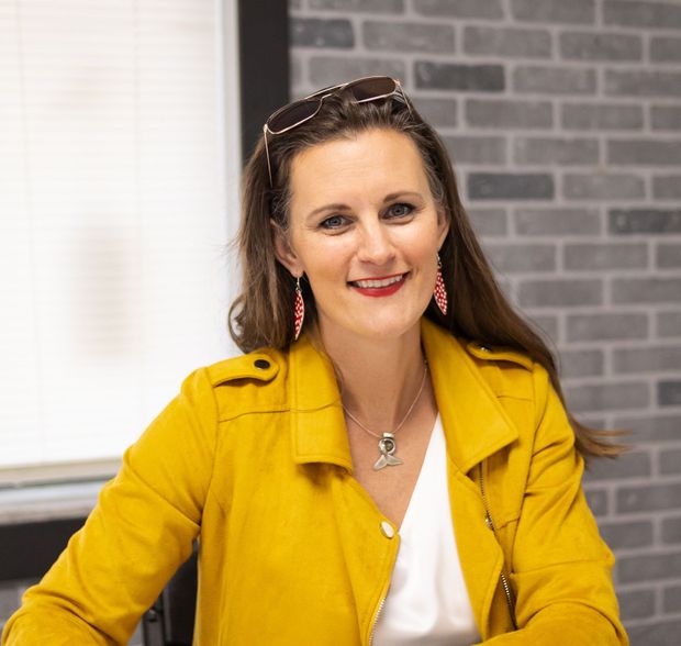 Woman with brown hair smiles, wearing a yellow jacket and red earrings, in front of a brick wall.