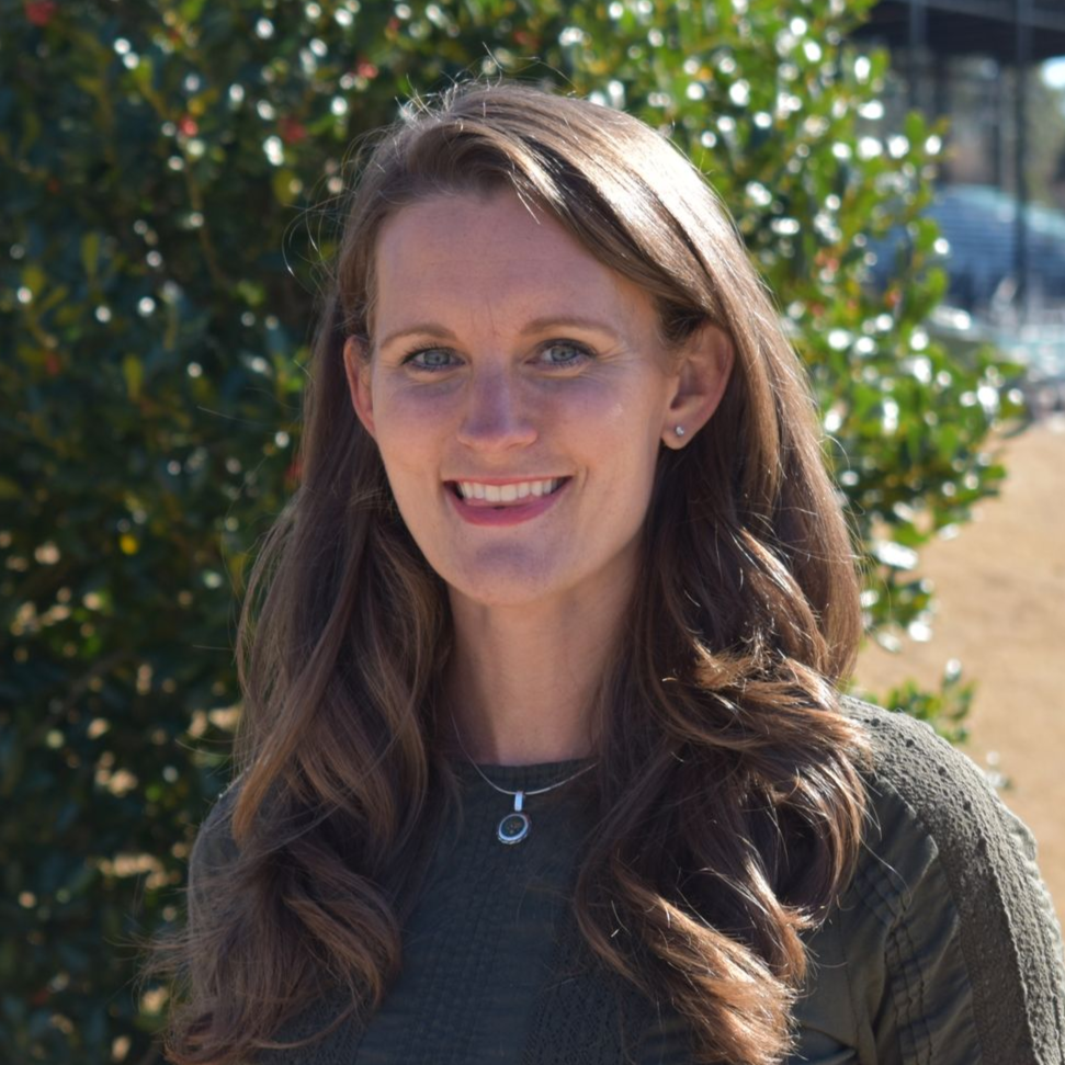 Woman with long brown hair smiling outdoors, wearing a green sweater and necklace.
