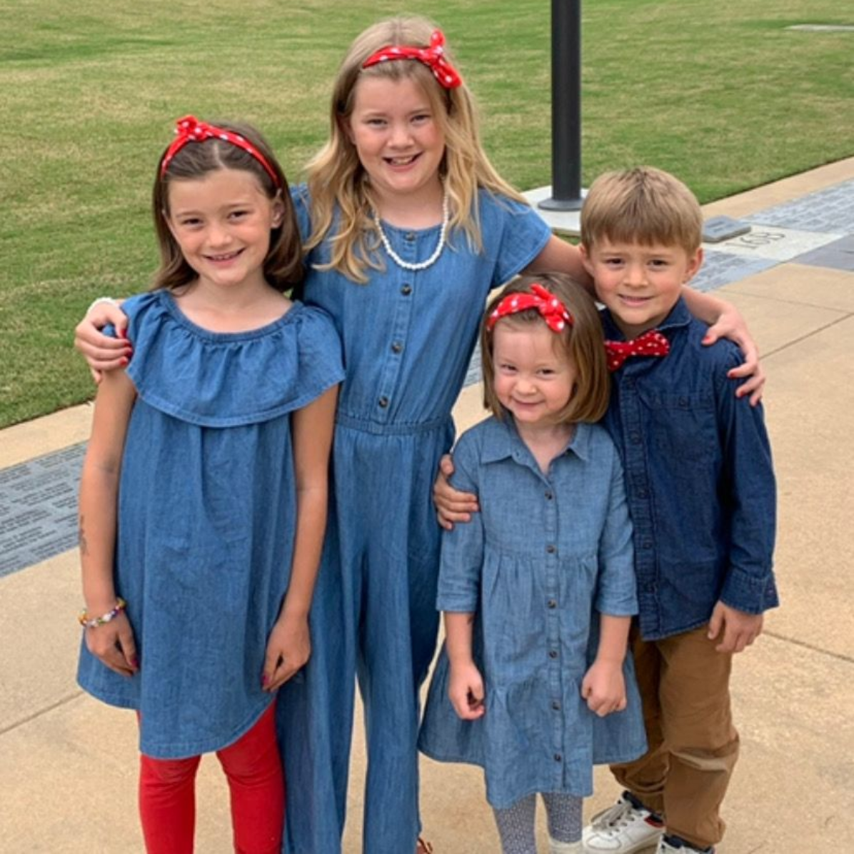 Four children smiling, wearing denim outfits with red accents, posing outdoors.