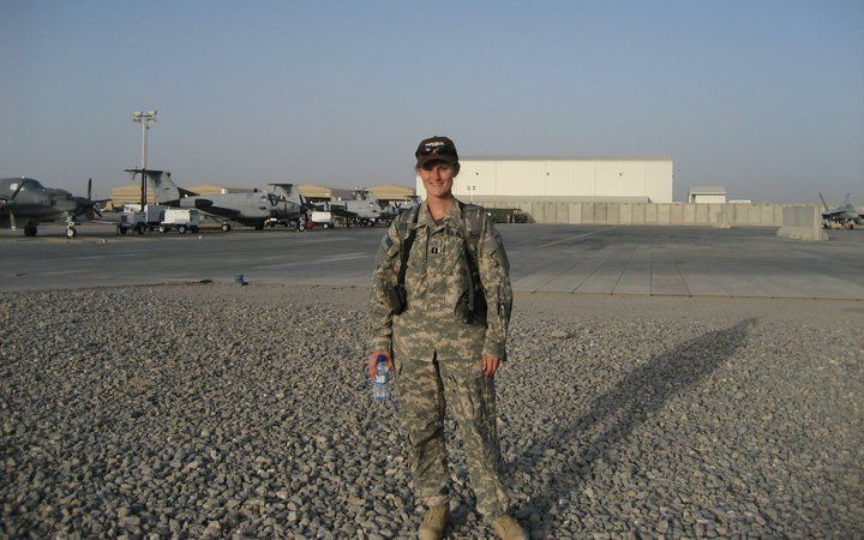 Soldier in camouflage uniform stands outdoors on gravel; airfield in background.