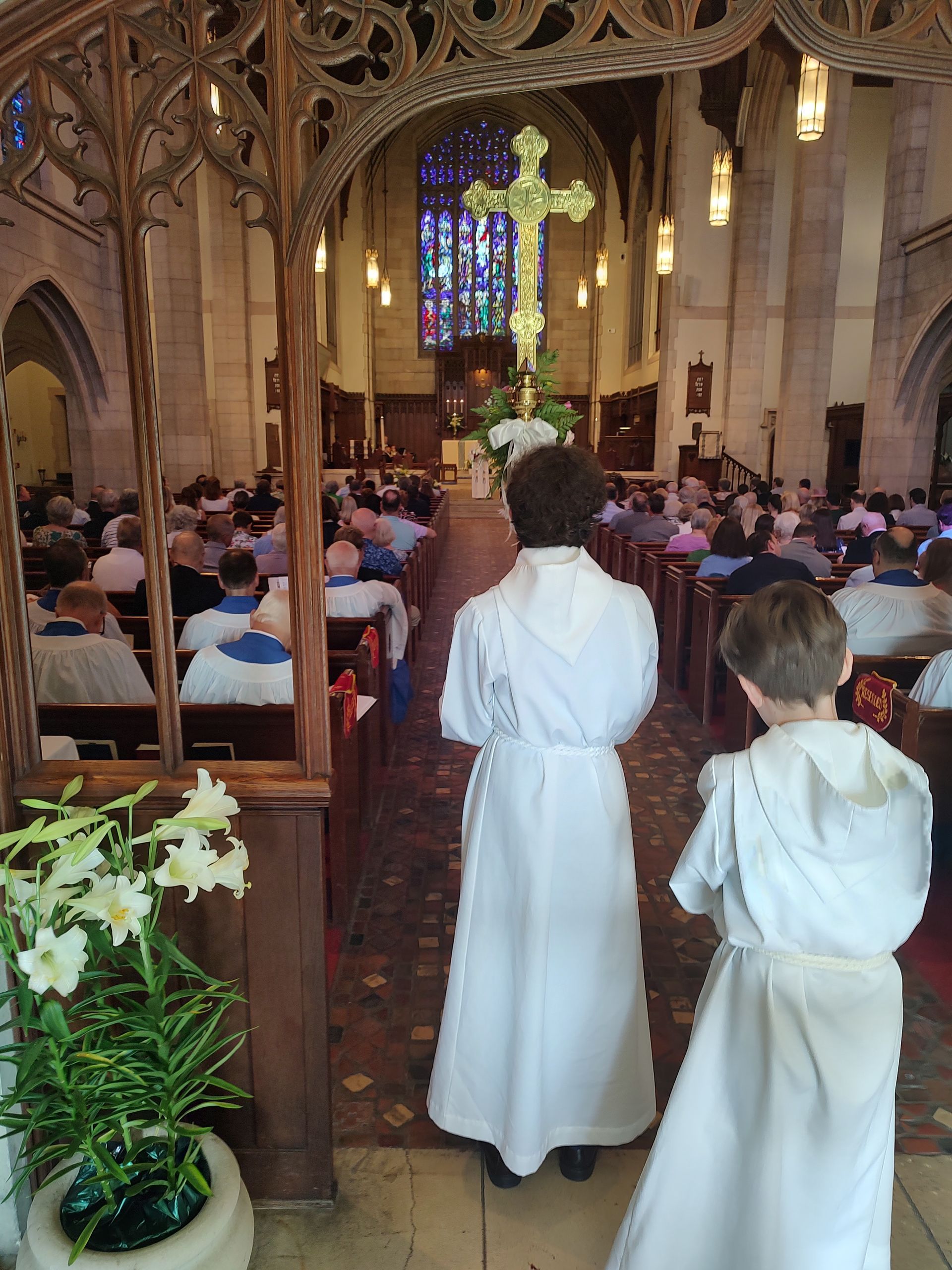 People in a church wearing white robes. Two youth processing. One carrying a cross and the other carrying a Bible. 