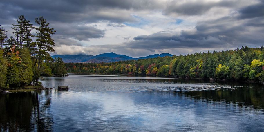 A calm lake reflects a cloudy sky and forested shoreline, with distant mountains under a dark sky.