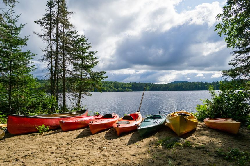 Colorful kayaks and a canoe on a sandy shore overlook a lake under a cloudy sky.