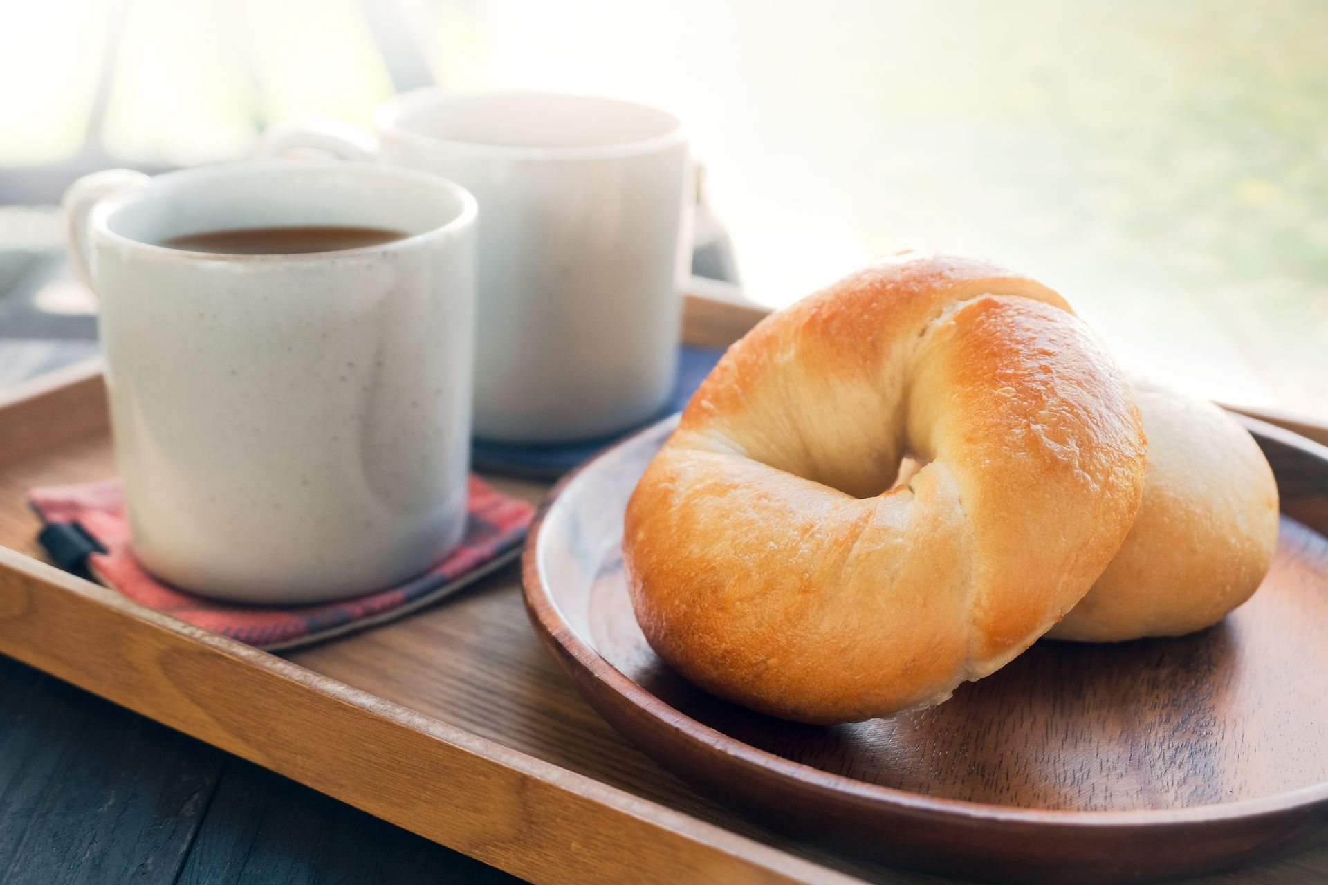 Coffee and bagels on a wooden tray, near a window. Two mugs of coffee, two bagels on a plate.
