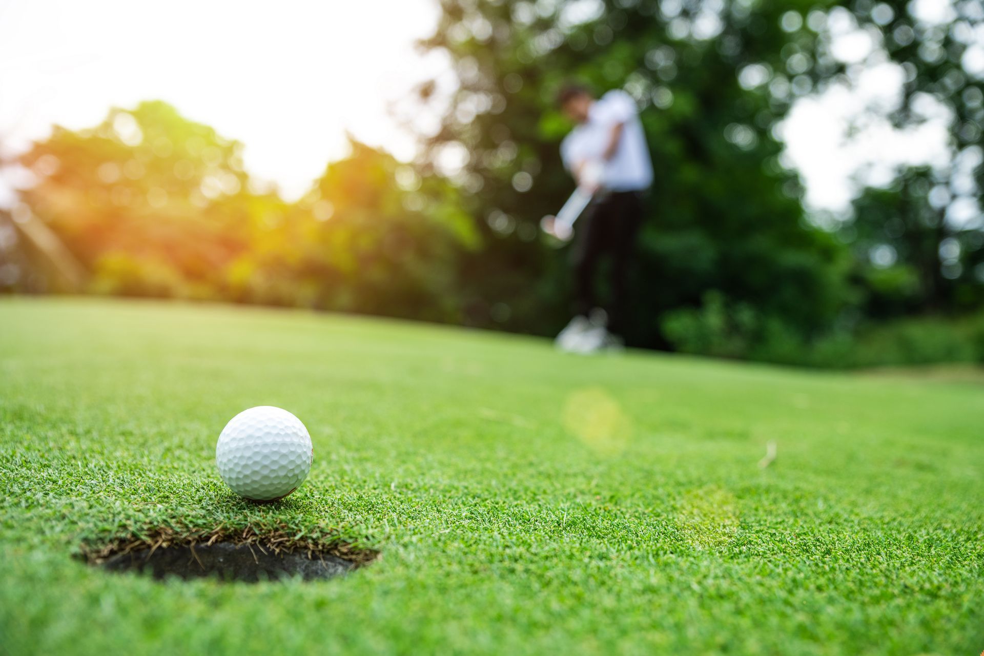 Golf ball near hole on green; golfer blurred in background about to putt.