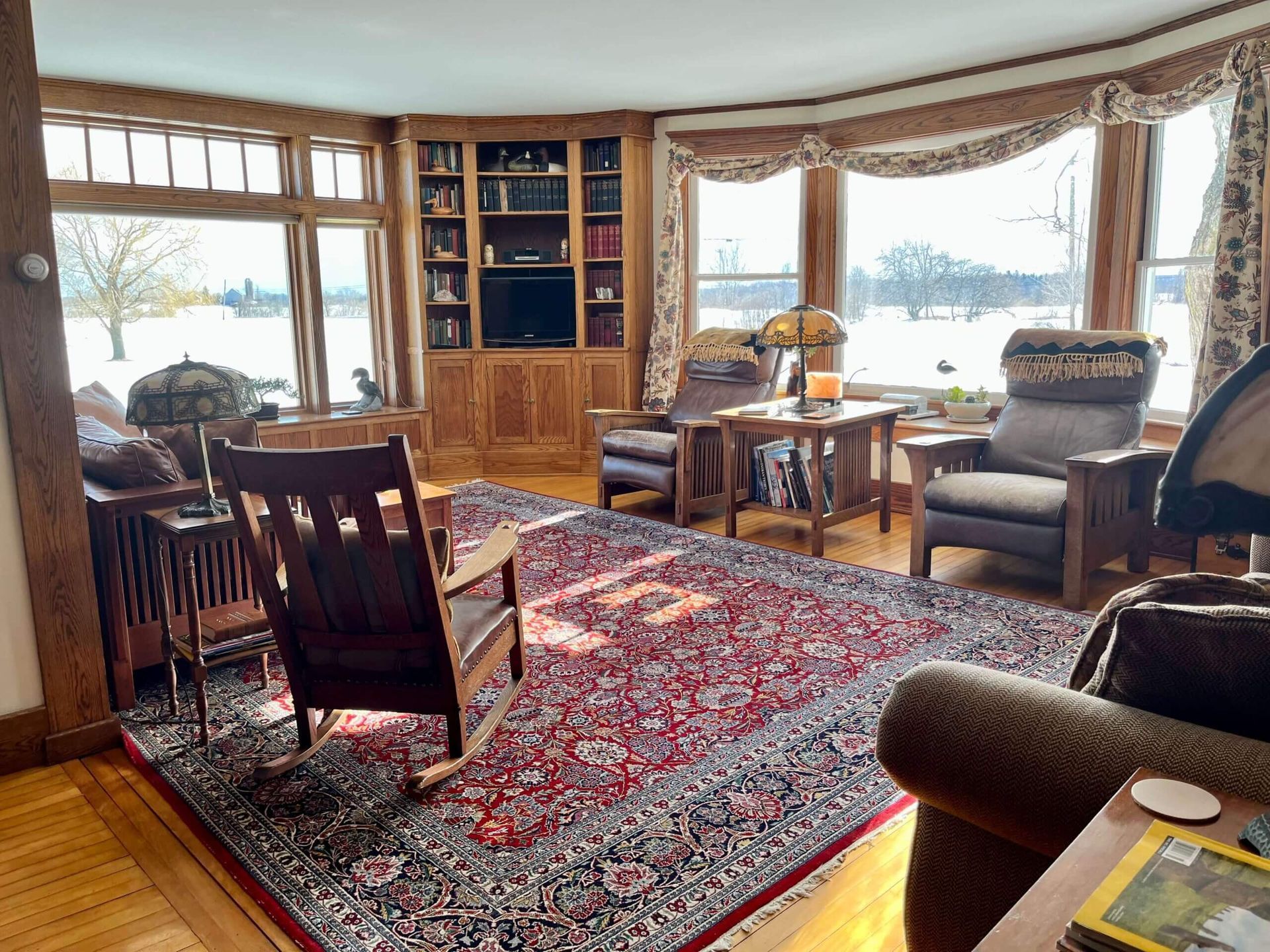 Living room with wood paneling, large windows, rug, rocking chair, and two armchairs; snowy outdoor view.