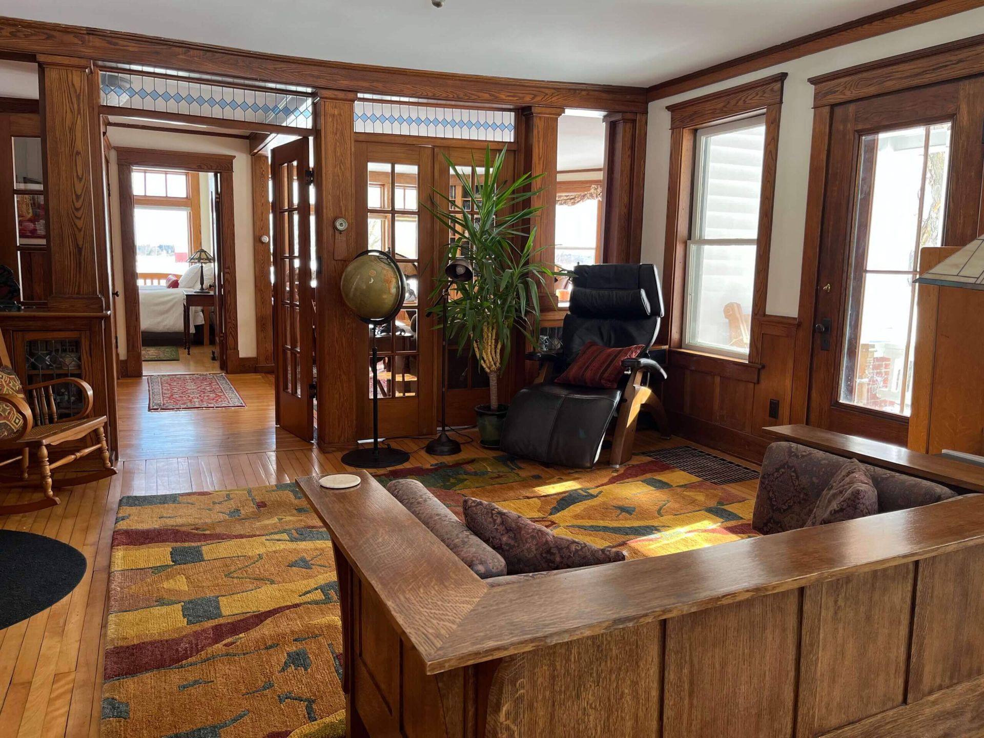 Warm-toned living room with wooden trim, sectional sofa, recliner, and decorative rug.  Sunlight streams through the windows.