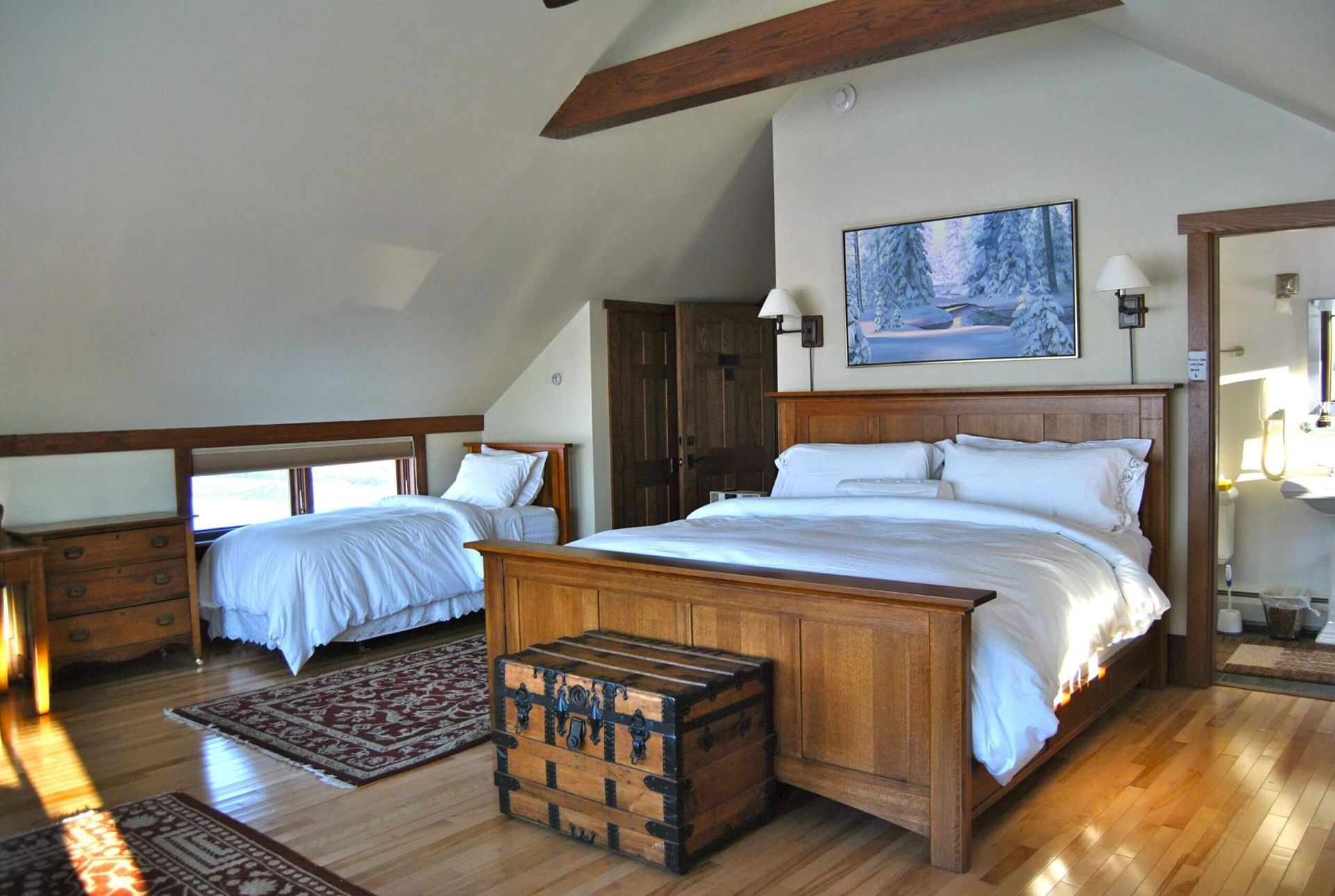 Bedroom with two beds, wooden furniture, and a trunk. Light-colored walls and hardwood floors.