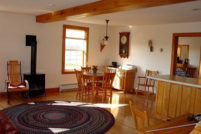 Bright, sunlit living room with wood floors, round rug, dining table, wood stove, and open doorway.