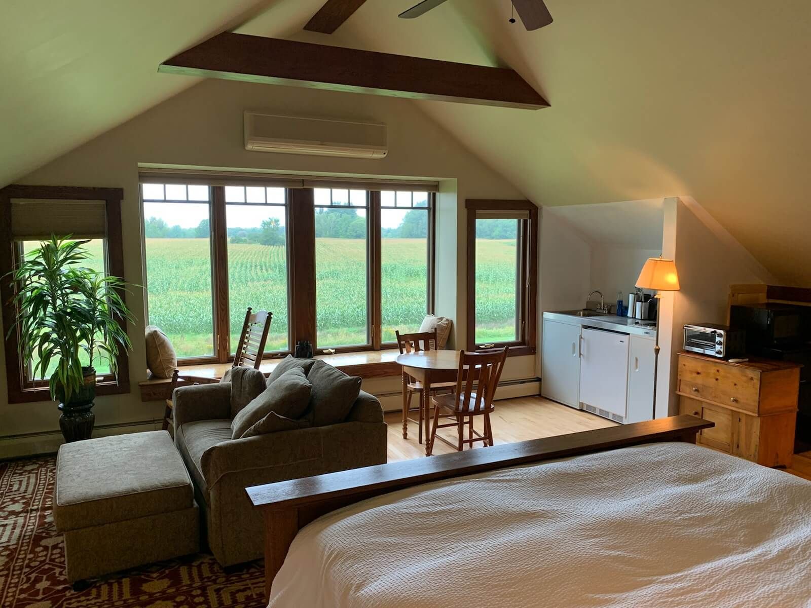 Cozy attic apartment with bed, couch, table, kitchenette, and large window overlooking a field.