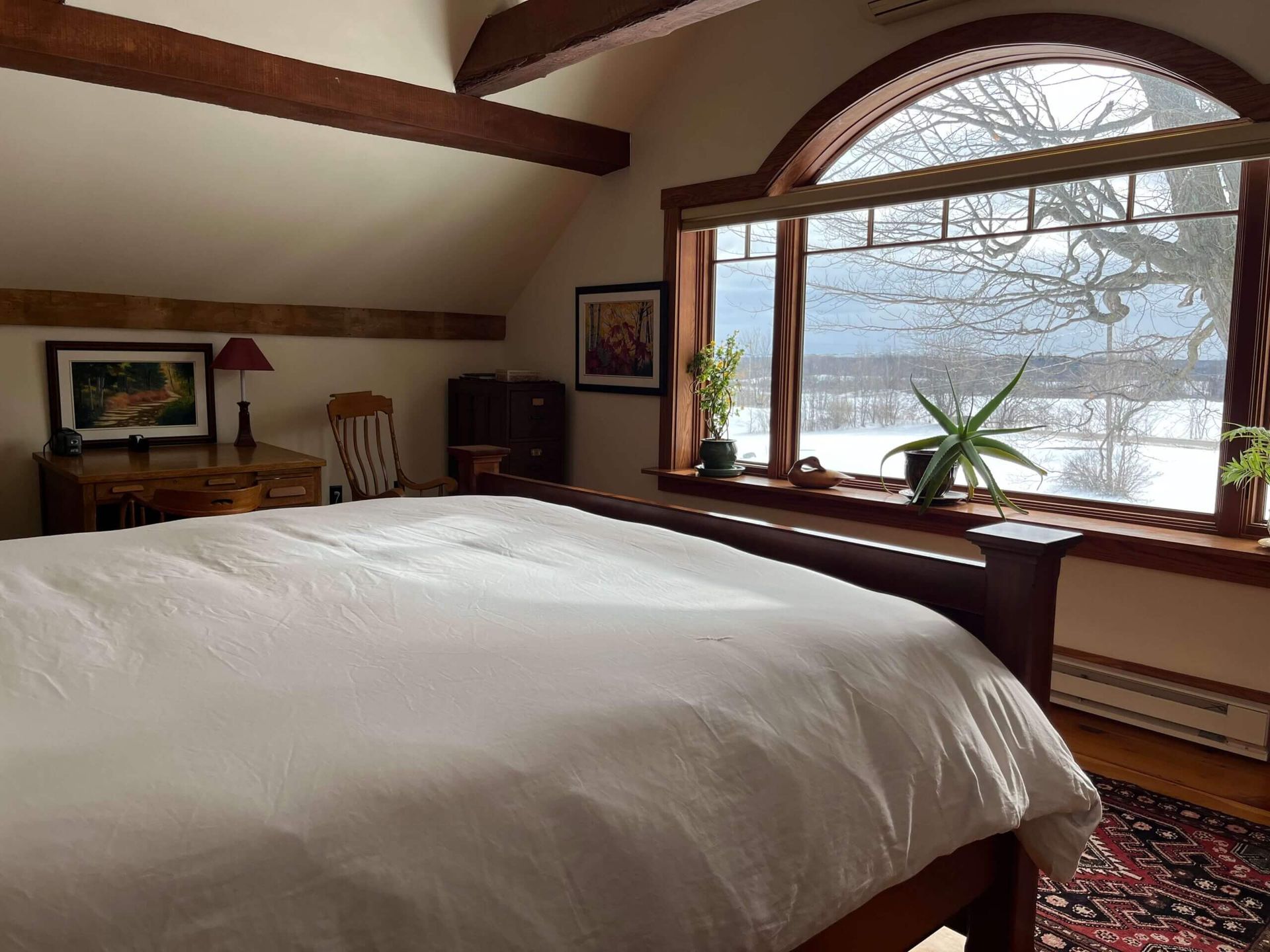 Bedroom with large arched window overlooking a snowy landscape. Bed, desk, and wooden beams visible.