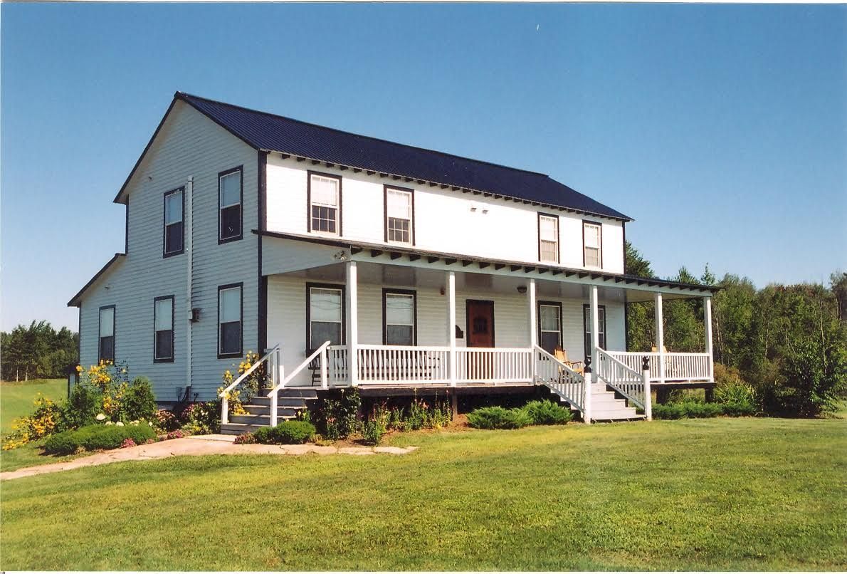 Two-story white house with a porch and black roof on a grassy field under a blue sky.