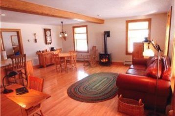 Spacious living room with hardwood floors, rug, wood stove, and leather sofa; a table and chairs are in the foreground.