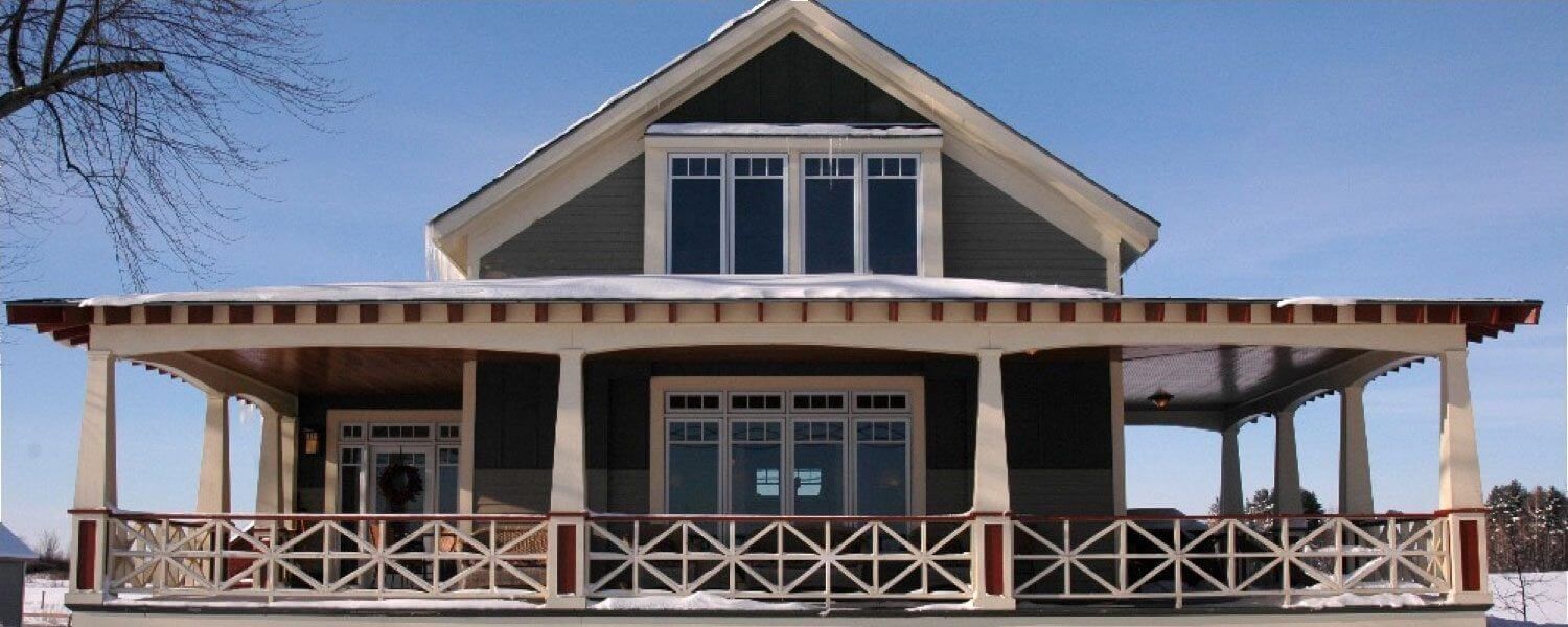 A two-story house with a large porch and decorative railing, covered in snow, on a clear, blue sky day.