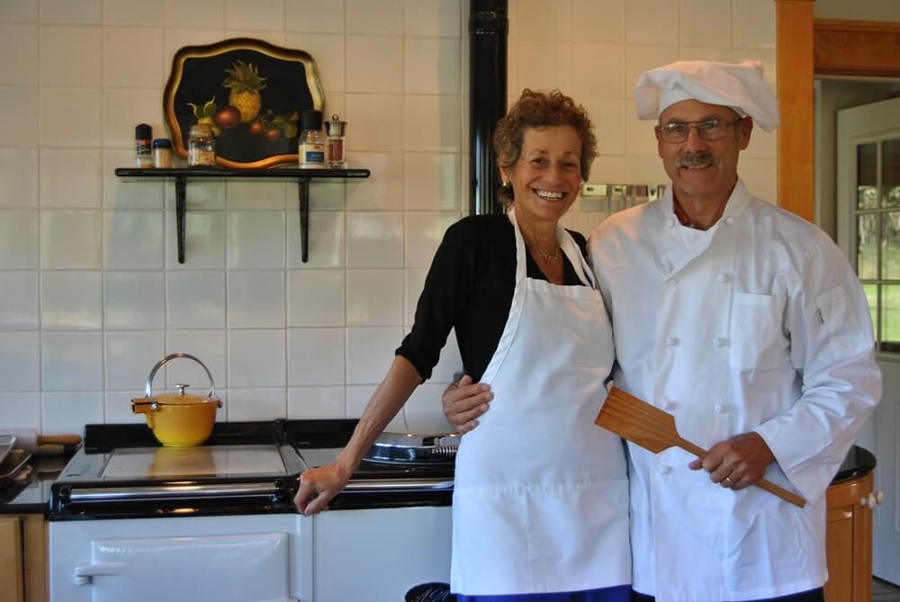 Couple in kitchen, one in chef's whites, smiling next to a white stove, holding a spatula.
