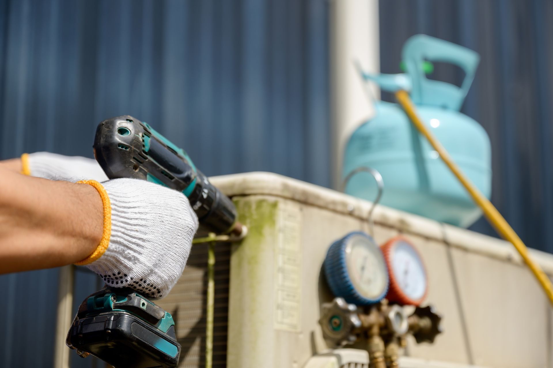 Person in gloves using a drill on an air conditioning unit; blue tank in the background.