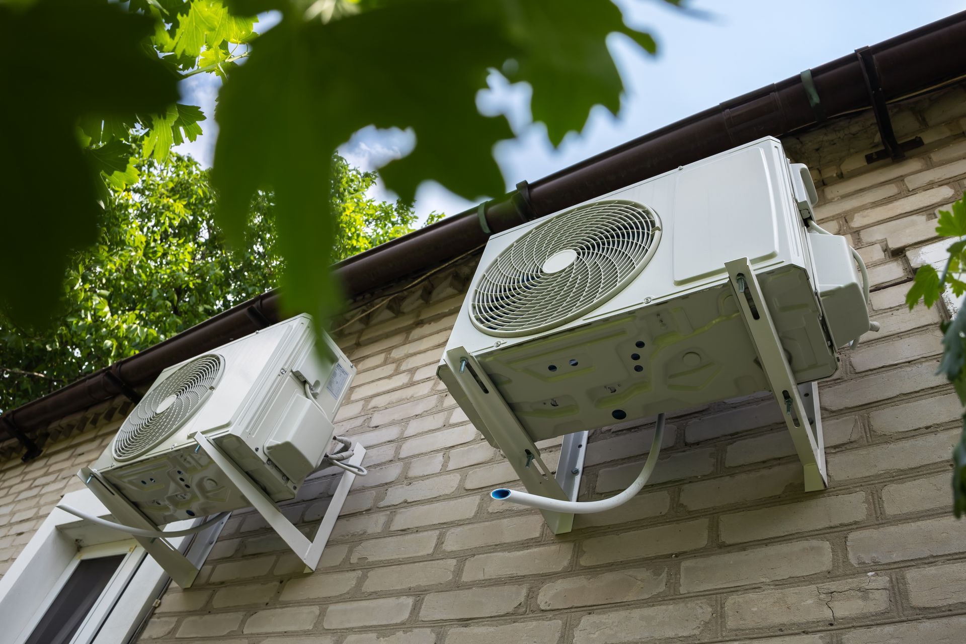 Two white air conditioning units mounted on a beige brick wall beneath green tree leaves. Two white air conditioning units mounted on a beige brick wall beneath green tree leaves.