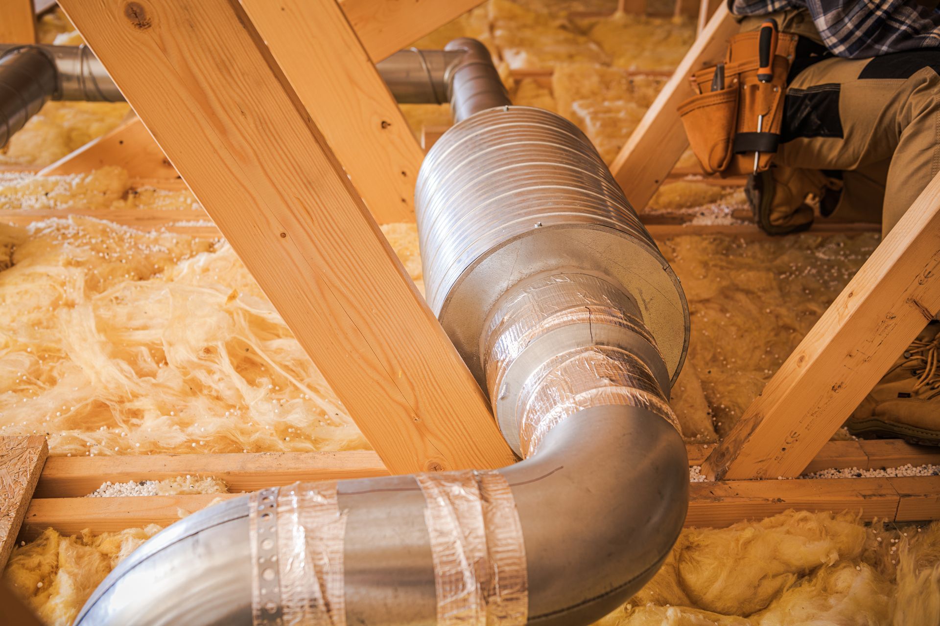HVAC ductwork installation in an attic, surrounded by insulation, with a worker partially visible.