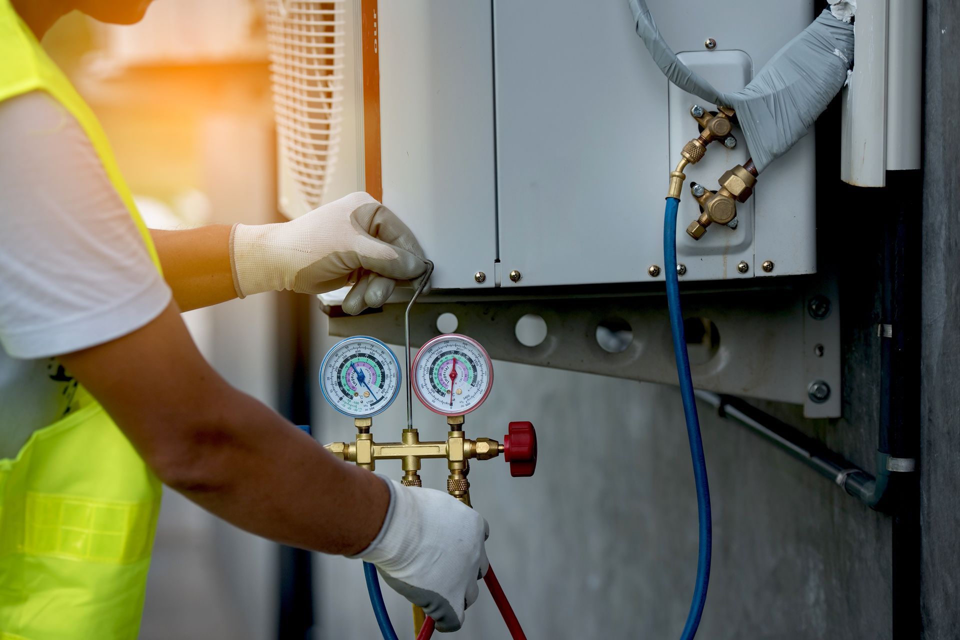 A technician wearing gloves and a safety vest uses a manifold gauge set to service an outdoor air conditioning unit.