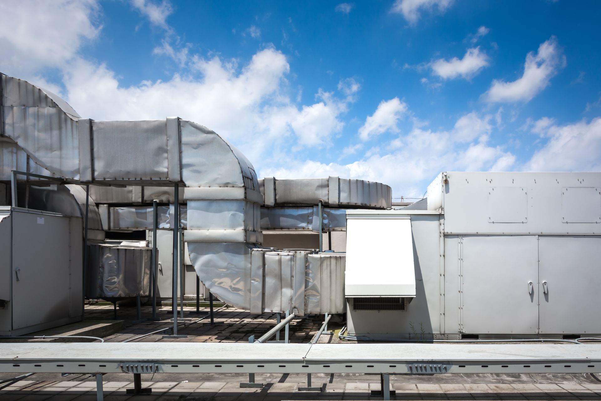 Rooftop HVAC units with metal ductwork, against a blue sky with fluffy white clouds.