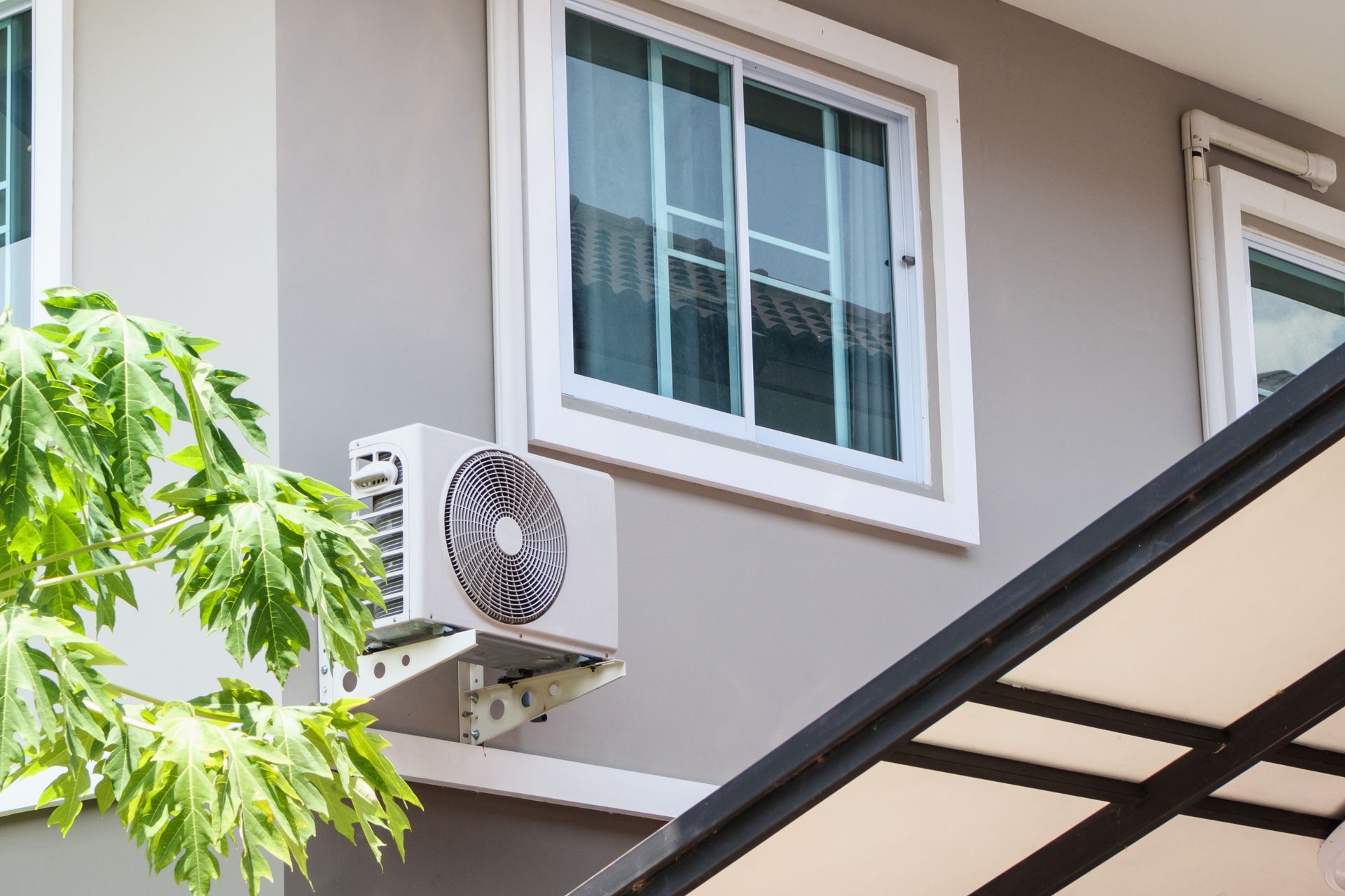 An outdoor air conditioning unit is mounted on a light gray exterior wall beneath a window, with a partial canopy nearby.