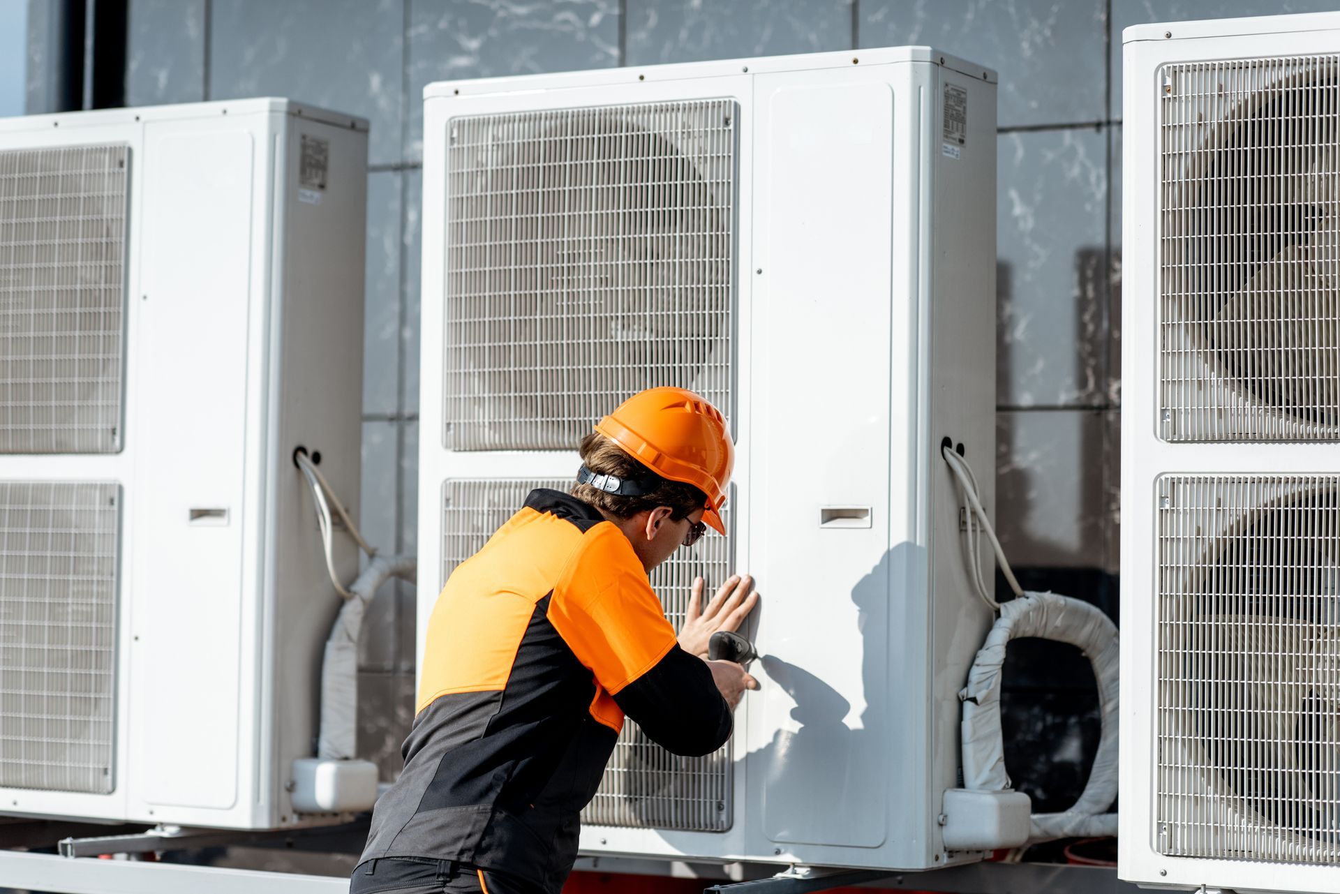 A technician in an orange hard hat and high-visibility jacket repairs an outdoor air conditioning unit.
