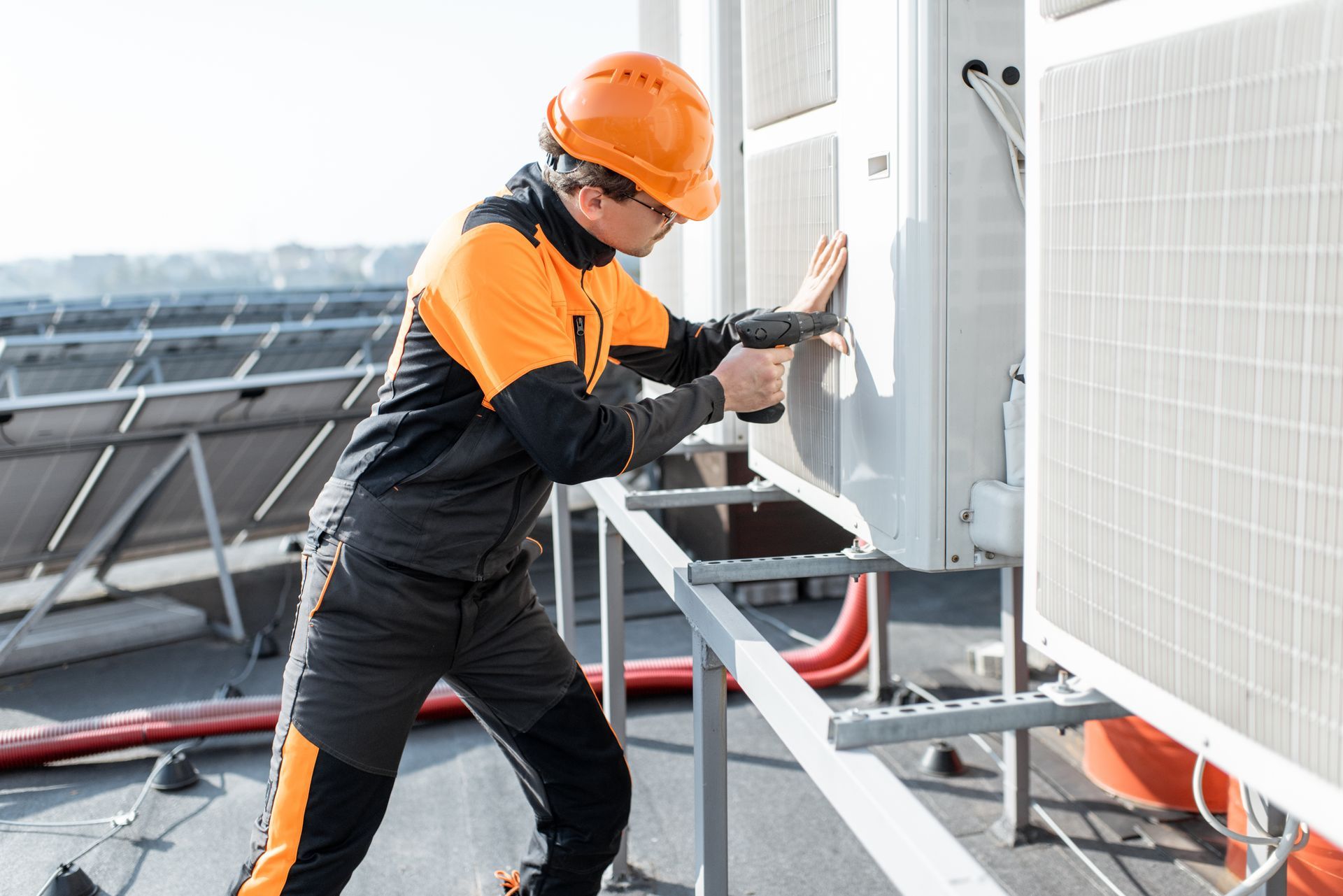 A worker in a high-visibility uniform and orange hard hat uses a drill on an outdoor HVAC unit on a rooftop. A worker in a high-visibility uniform and orange hard hat uses a drill on an outdoor HVAC unit on a rooftop.