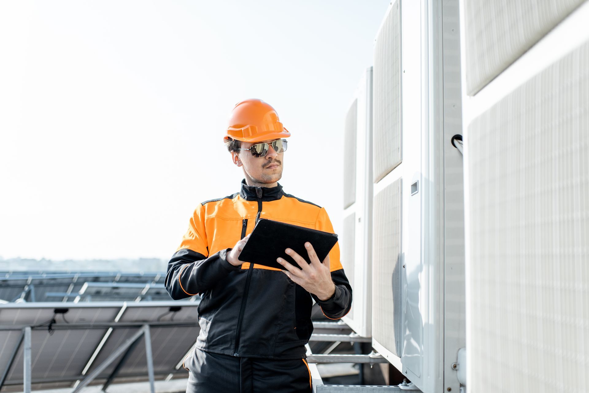 Technician in a hard hat and high-visibility jacket using a tablet to inspect HVAC units on a building roof.