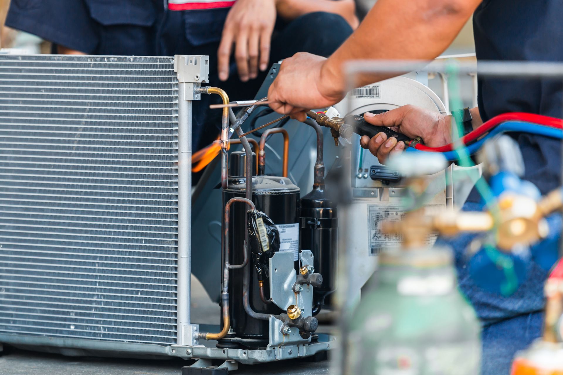 Hands of a person repairing an air conditioning unit; other person visible in background.