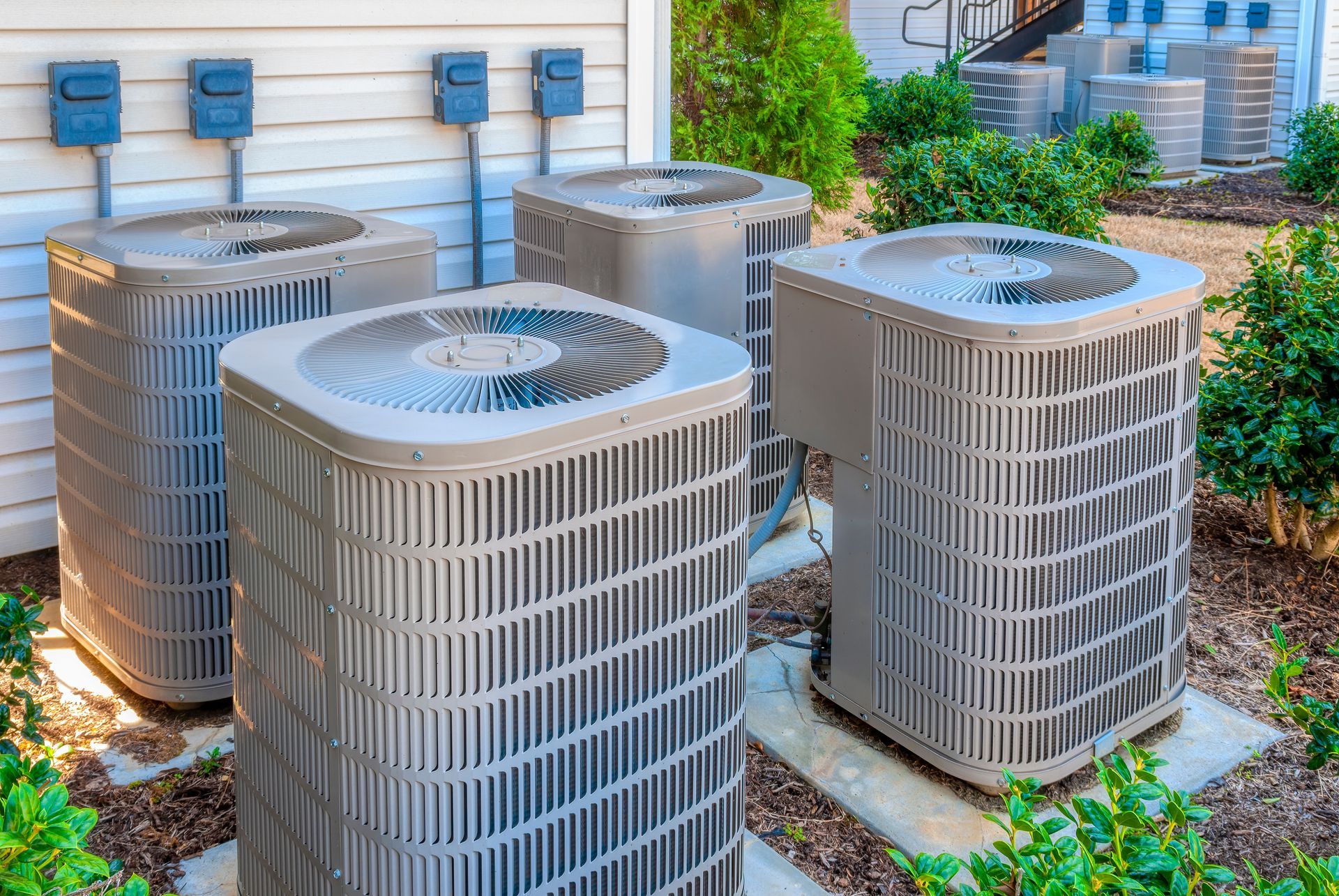 Four outdoor air conditioning units near a building, surrounded by greenery.
