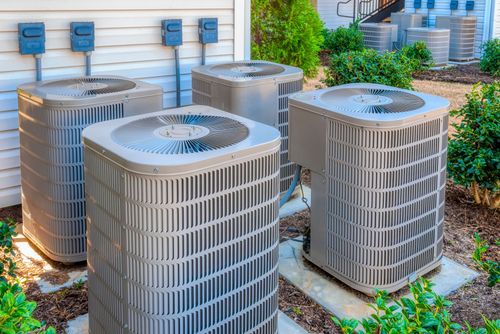Four outdoor air conditioning units near a building, surrounded by greenery.