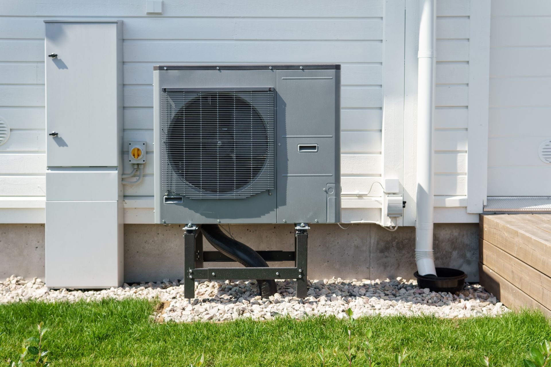 Exterior air conditioning unit mounted on a white house, with electrical box and drainage pipe.